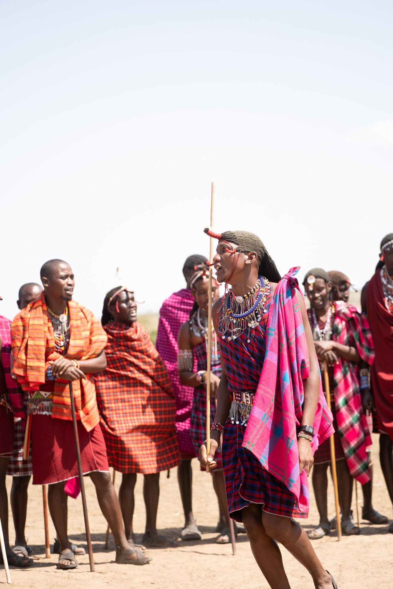 Maasai men in beaded jewellery and bright plaid shuka cloth gathered for a community greeting, one striding forward with a herding staff as the rest of the group looks on.